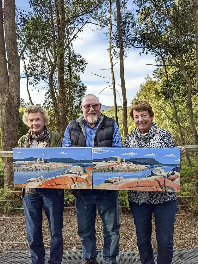 Two sisters aholding their paintings re standing either side of artist, John Gibb. The background is a bush and mountain scene.