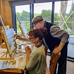 A male artist is assisting a woman with her painting.