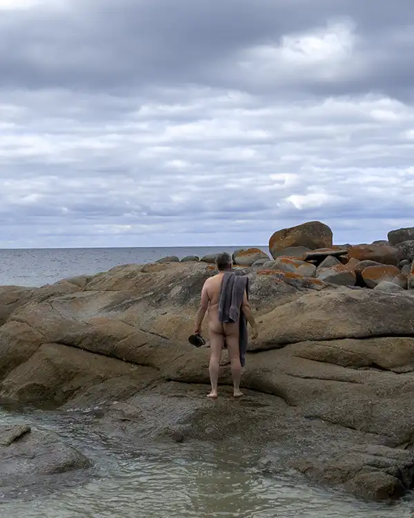 Graham is facing away from the camera. He is standing on a rock slab at the beach, looking out to the ocean. He is not wearing colthes, but has a towel over his shoulder.